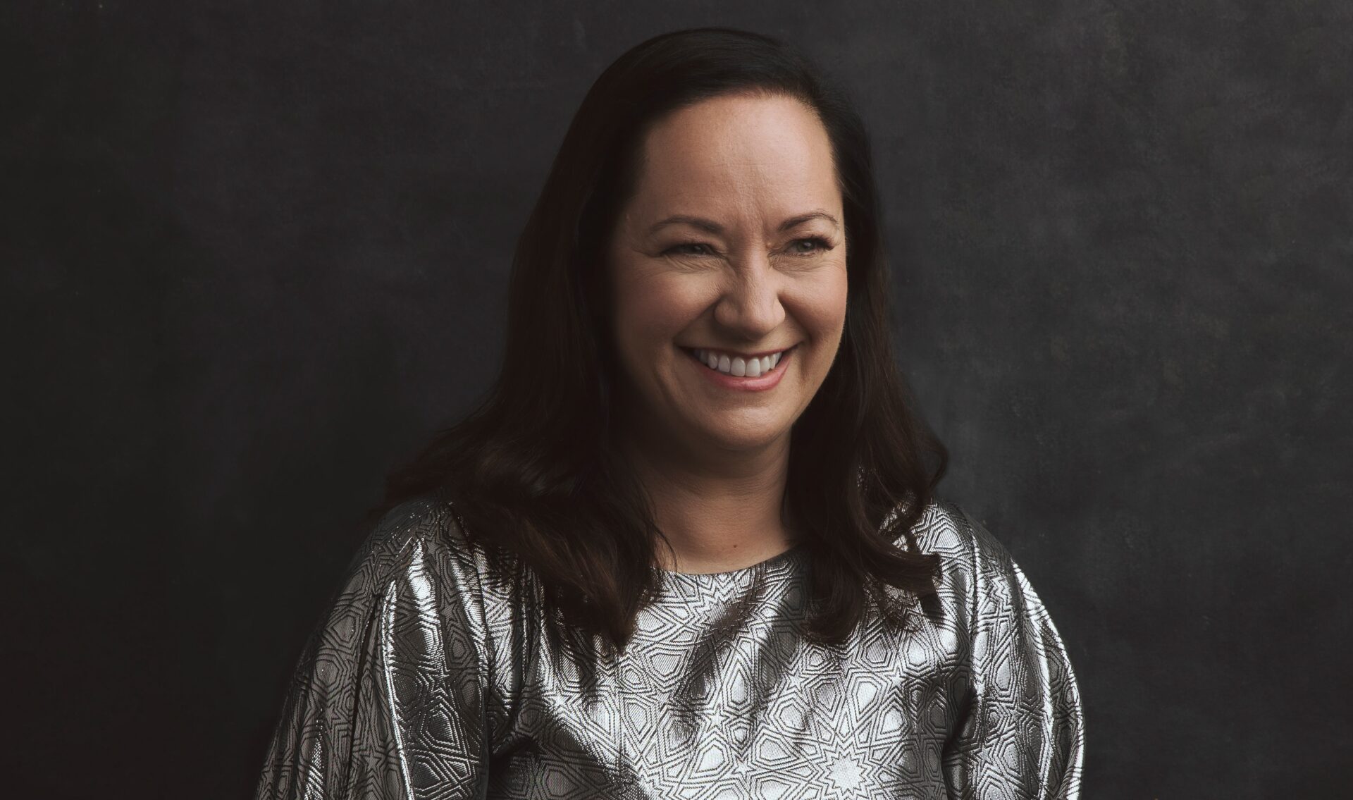 White woman with shoulder length black hair wearing a silver top - she is smiling