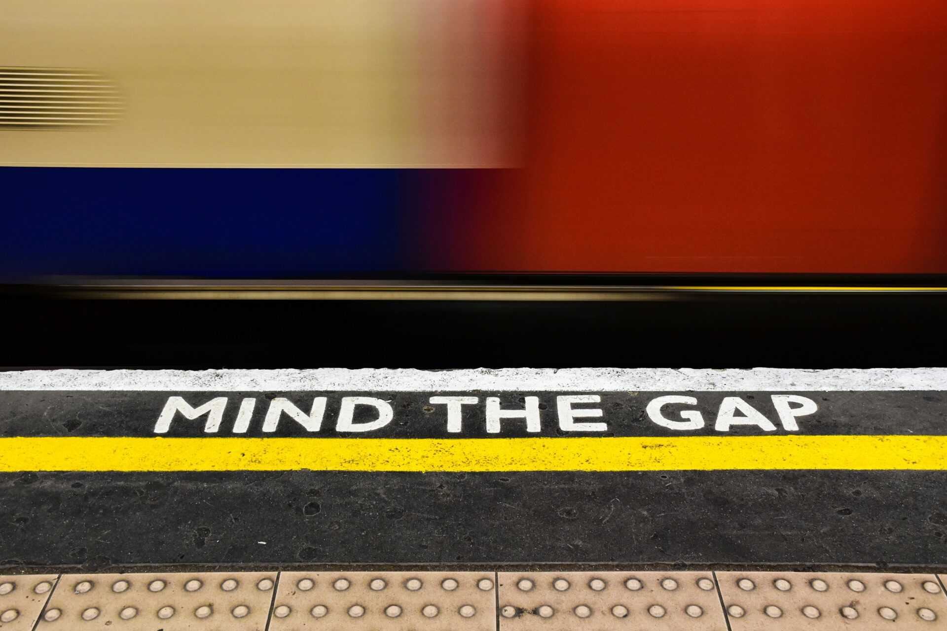 Photo of a train station platform close up with 'Mind The Gap' written on it and a train whizzing by