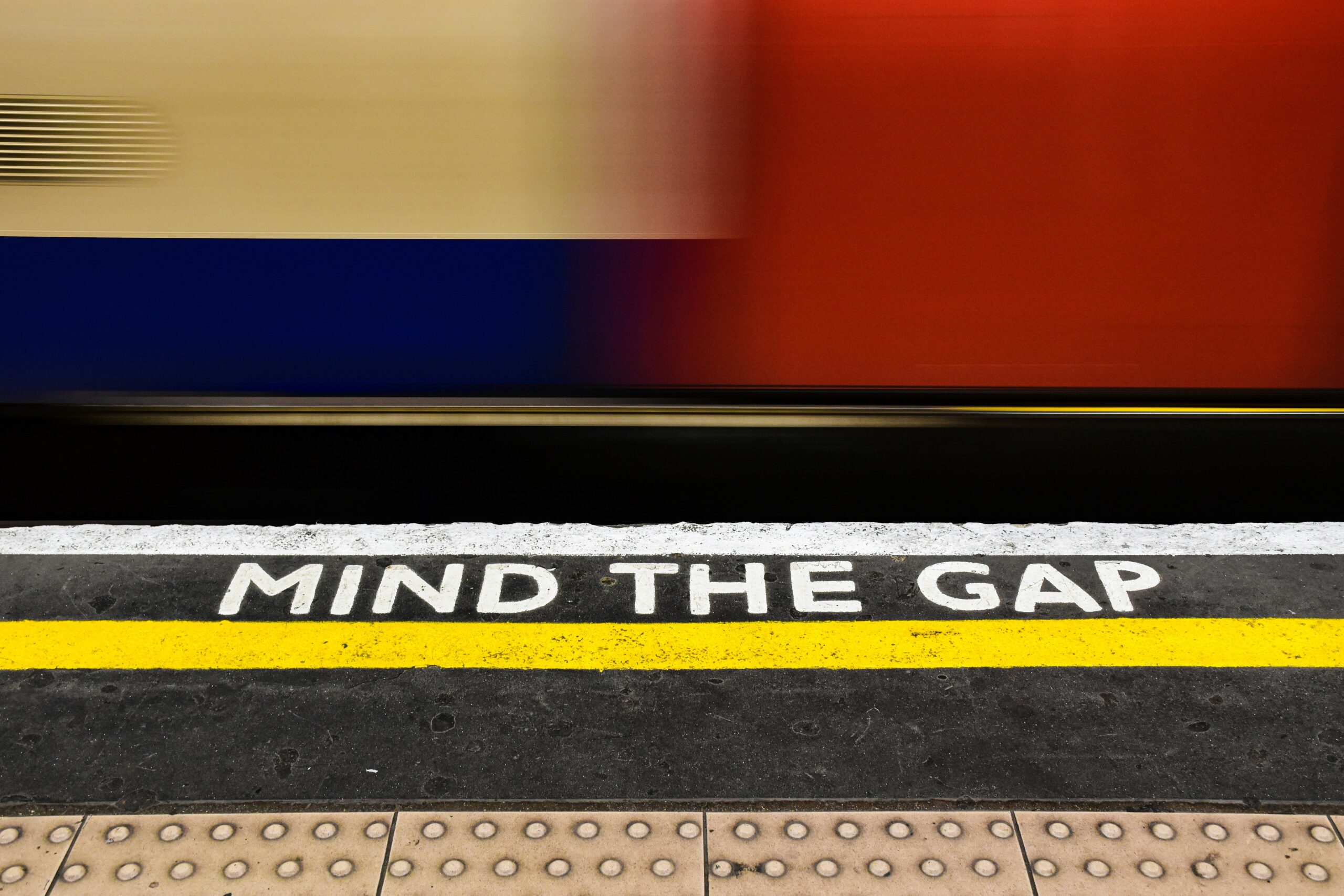 Photo of a train station platform close up with 'Mind The Gap' written on it and a train whizzing by