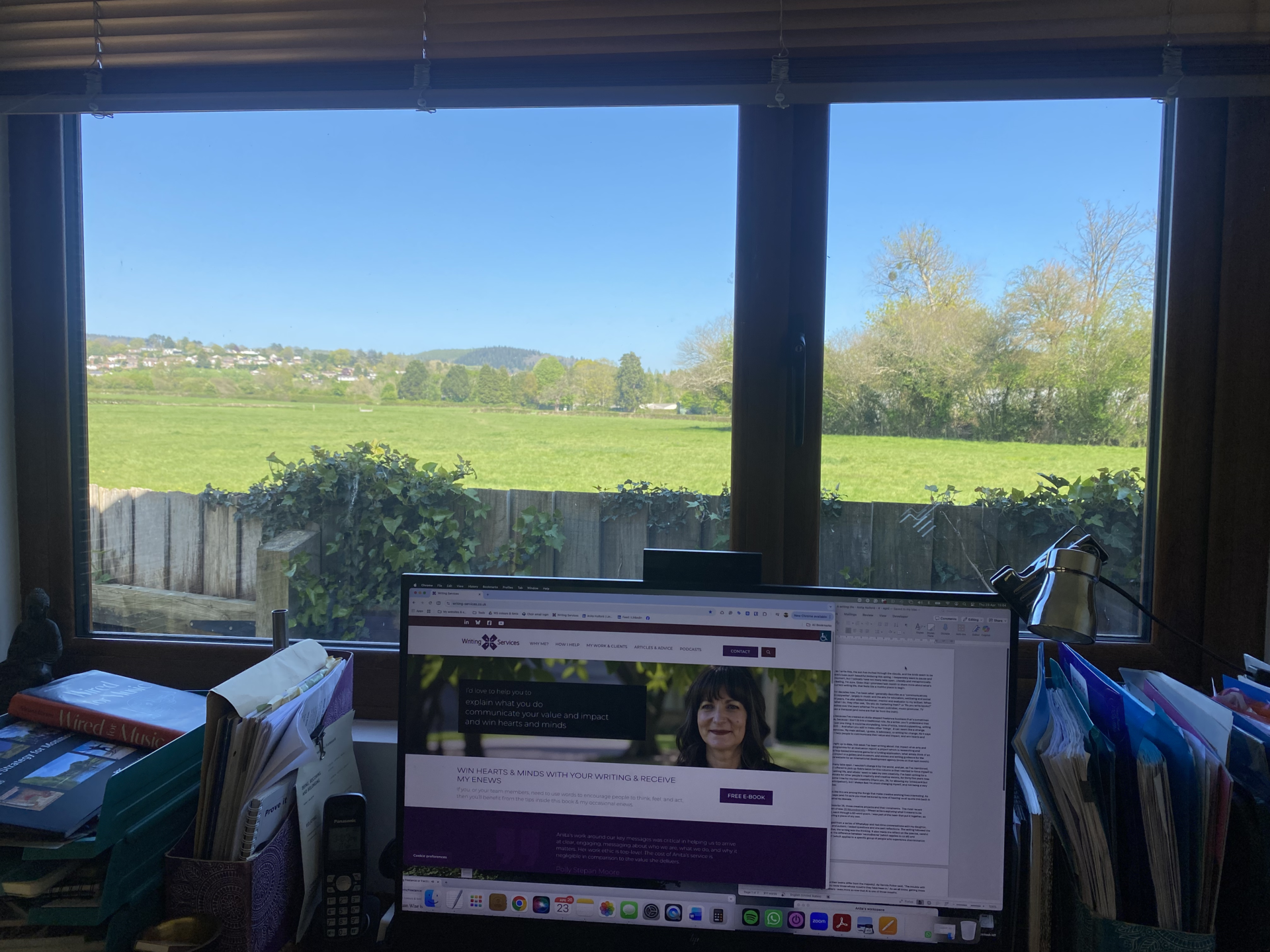 A computer display featuring Anita's website, below which is her desk, with files and books either side, and behind all of this is a window looking out onto a field, with blue sky above