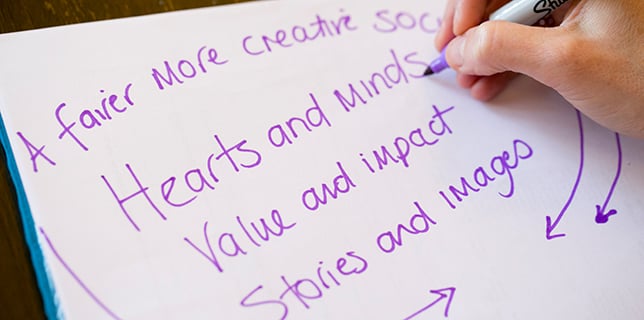 Fingers on a pen, writing the following words which are purple on a white sheet of paper: A fairer more creative society, hearts and minds, value and impact, stories and images